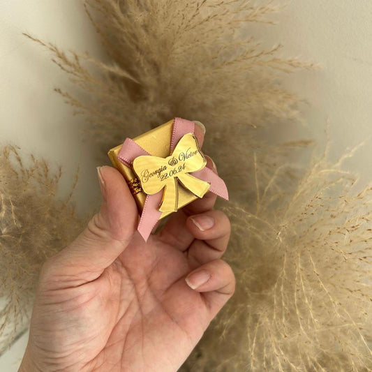 Boîte de dragées pour mariage, coloris doré avec ruban rose, personnalisée avec prénoms et date sur un nœud miroir
