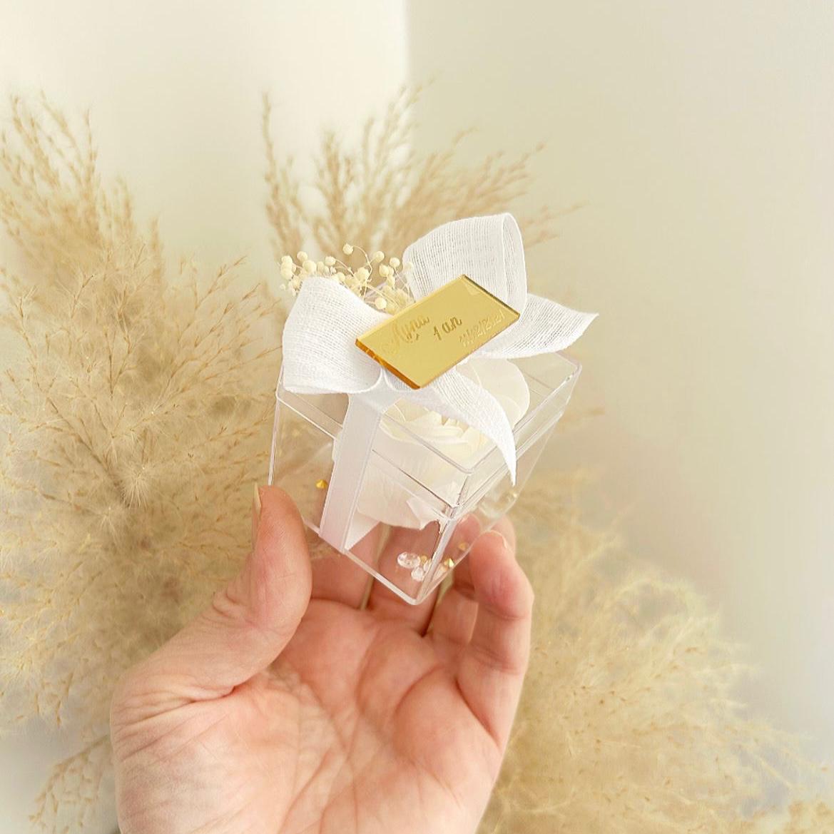 Lot de roses de savon Angelita en boîtes transparentes, décorées de gypsophile, avec nœud en ruban blanc et plaque dorée personnalisée, présentées sur un plateau.